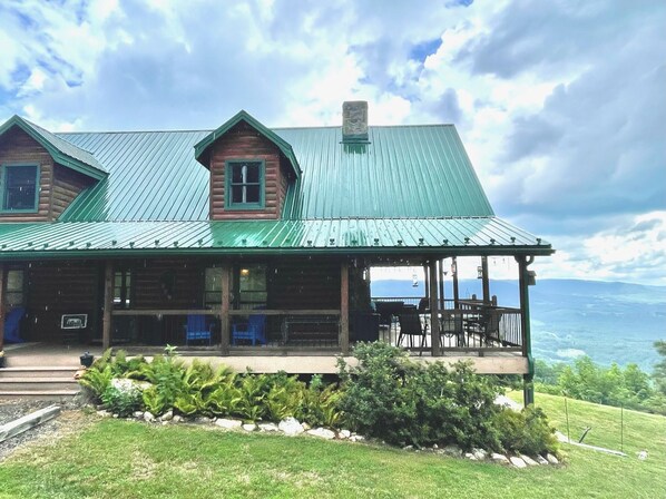A log cabin with a green metal roof, a porch with blue chairs, and a view of the mountains.
