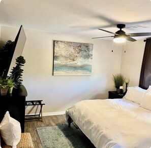 A bedroom with a bed, a ceiling fan, a painting, a nightstand, and a plant.
