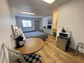 A compact hotel room with two single beds, a small kitchenette, a dining area with a table and chairs, and a television.