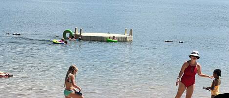 A lakeside scene with people in swimwear, a dock, and a grassy area.