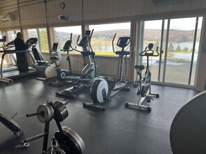A gym with various exercise equipment, large windows with a view of a lake and hills, and a person using a treadmill.