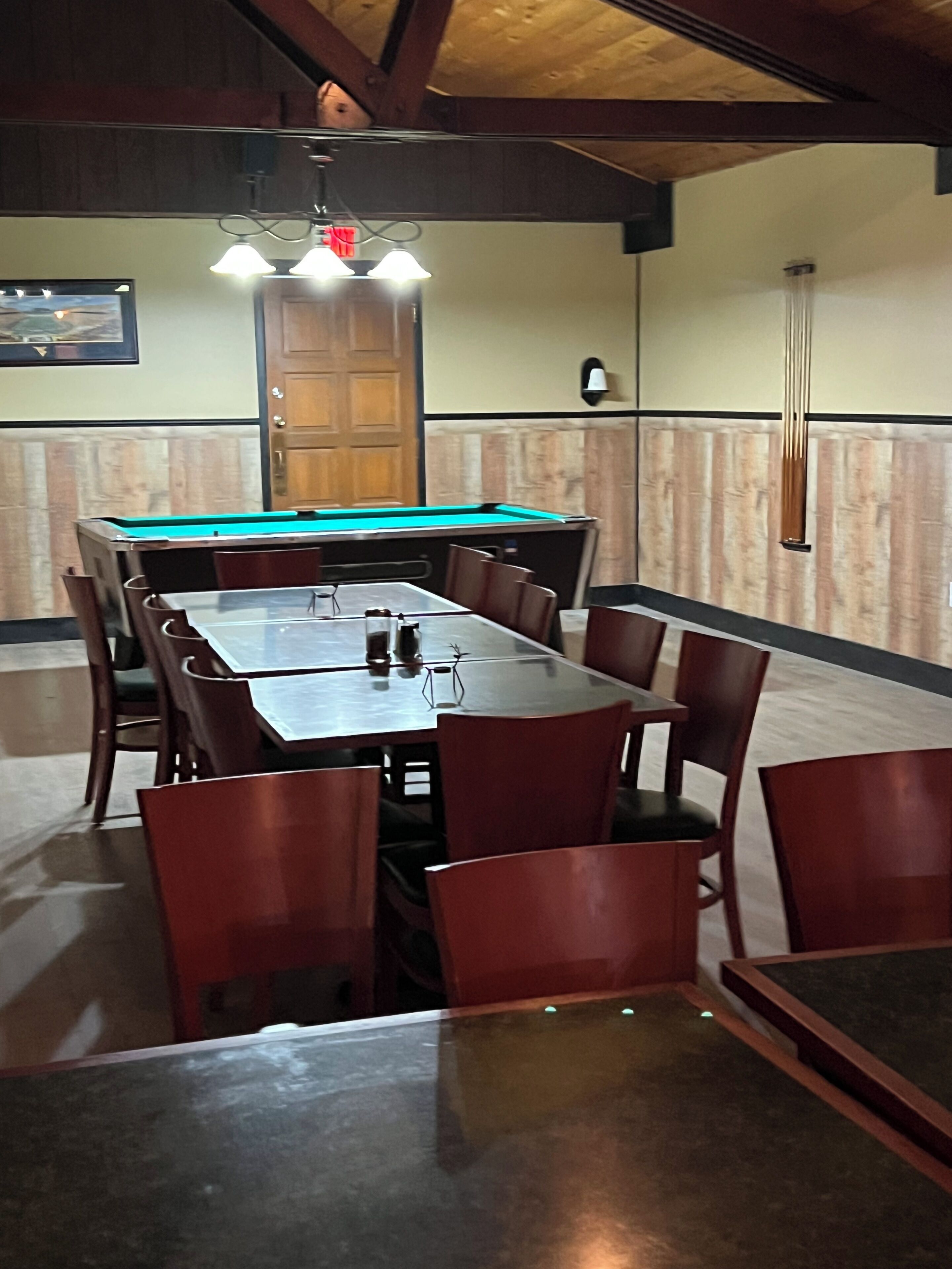 Una mesa de billar rodeada de sillas, techo de madera y una puerta al fondo.