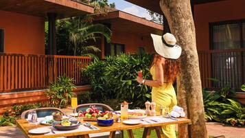 Mesa de comedor al aire libre con comida y bebidas, rodeada de follaje y un edificio con balcones de madera.