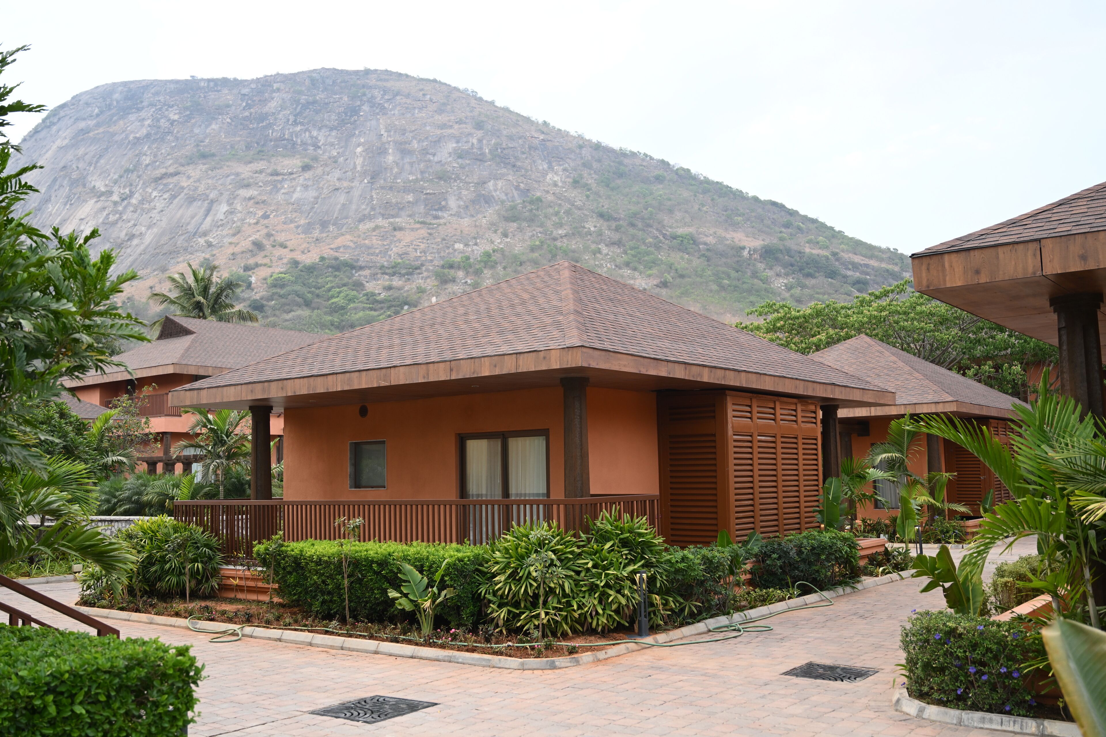 Un hôtel situé au pied d’une montagne, avec un bâtiment principal doté d’un toit en tuiles, entouré d’une végétation luxuriante et d’un chemin pavé.