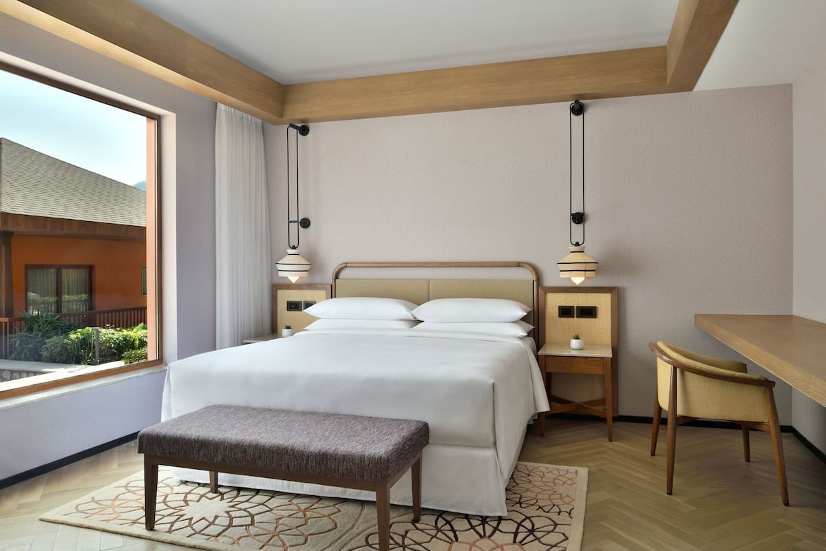 Habitación de hotel con una cama grande, un escritorio y un banco. Una ventana ofrece vistas al exterior.