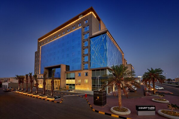 A modern hotel with a glass facade, a courtyard with palm trees, and a sign reading "COURTYARD BY HARBOR". - Courtyard By Marriott Jubail (Al Jubail)