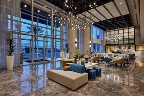 A spacious lobby with a modern design, featuring a large seating area, a kitchen in the background, and large windows providing a view of the outside. - Courtyard By Marriott Jubail (Al Jubail)