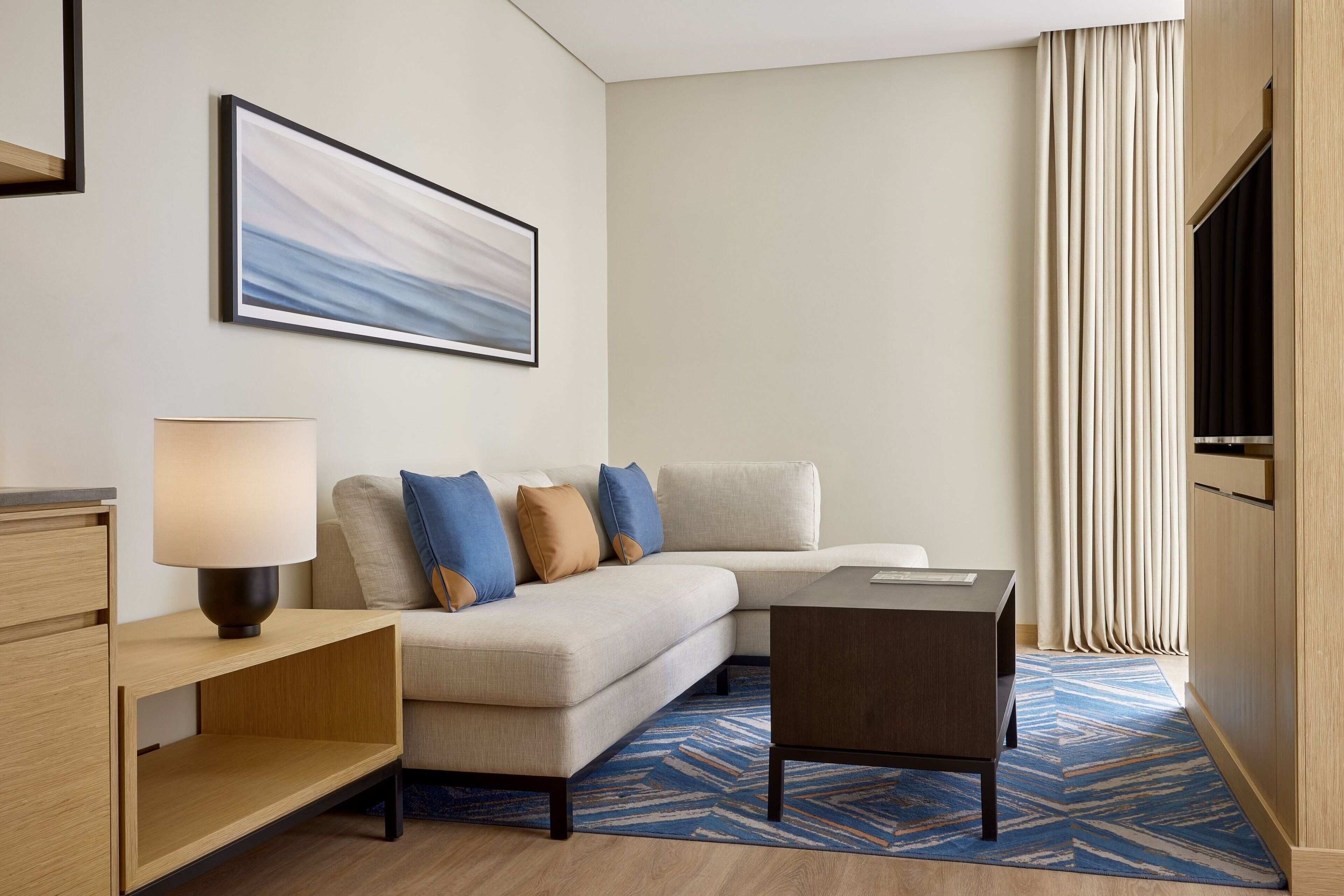 A modern hotel room with a sofa, a small table, a lamp, a TV, and a large window with curtains.