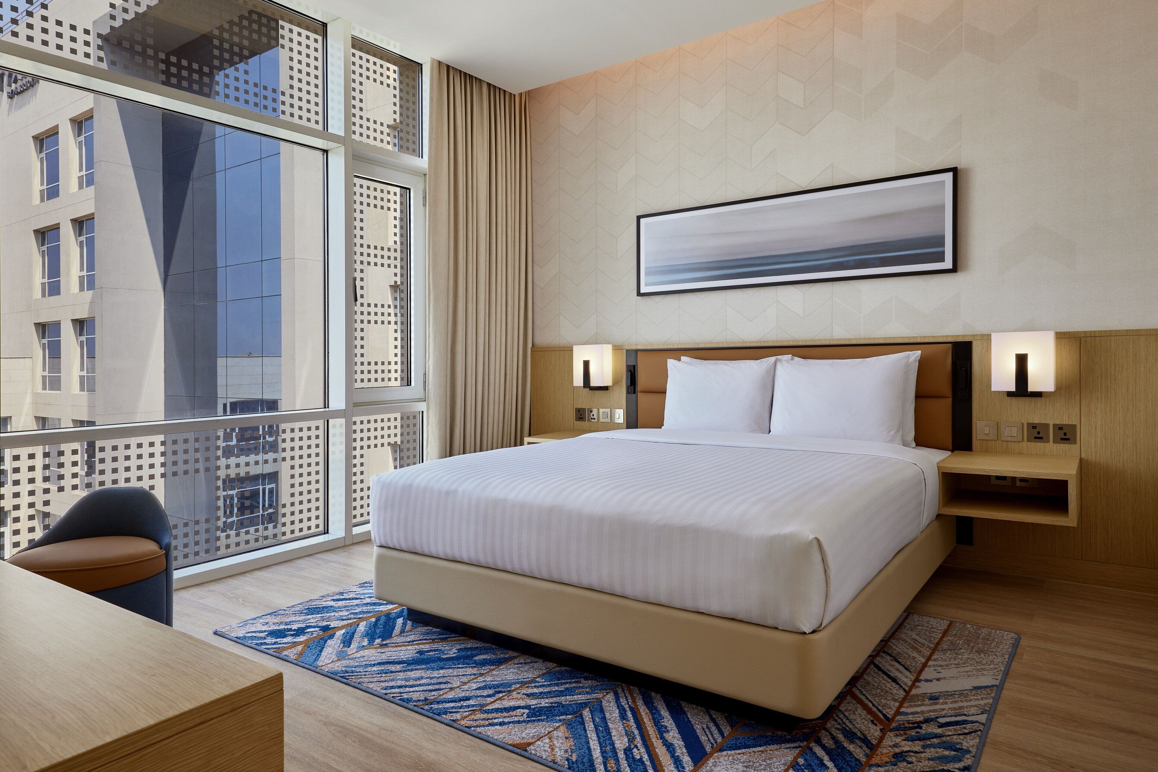 Una habitación de hotel moderna con una cama grande, un escritorio y vistas a la ciudad a través de un ventanal.