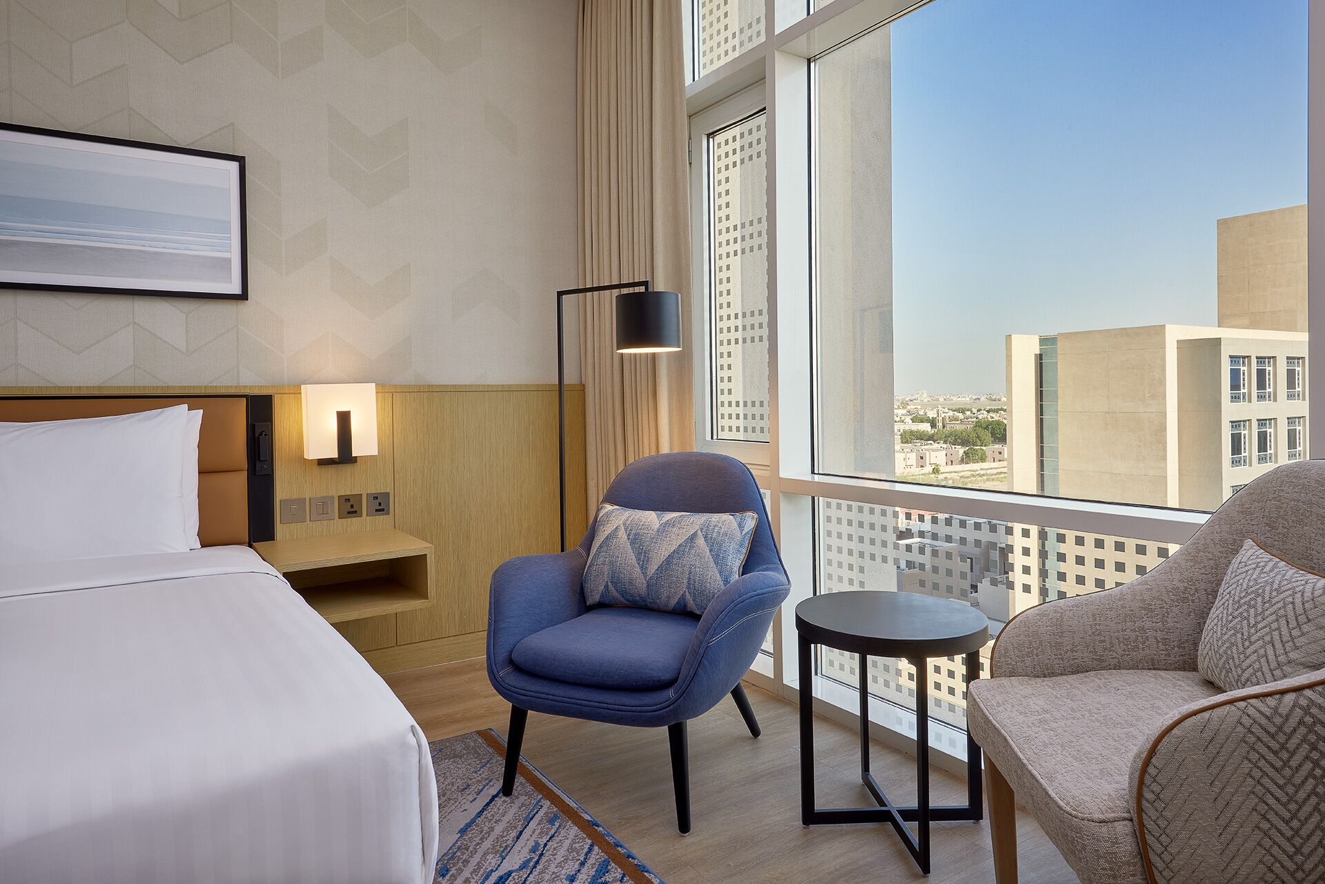 A hotel room with a bed, a blue armchair, a beige armchair, a small table, and a large window offering a city view.
