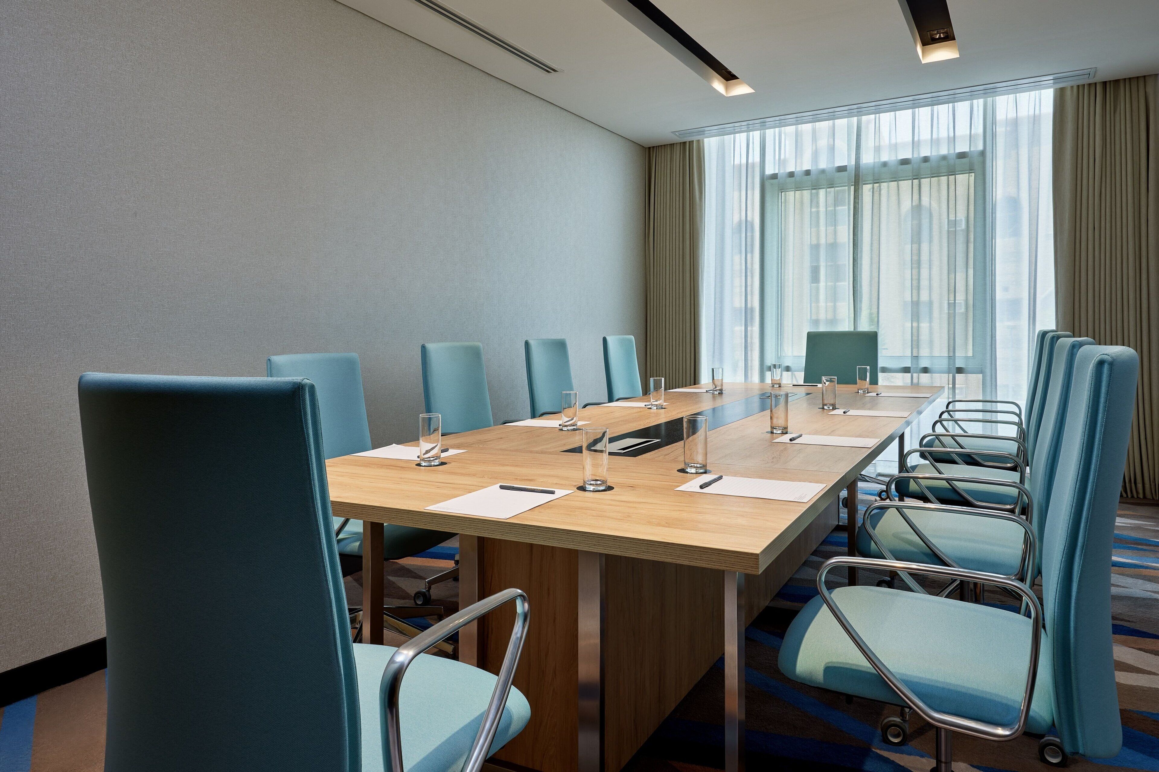 Une salle de conférence avec une longue table, des chaises bleues et une grande fenêtre avec des rideaux.