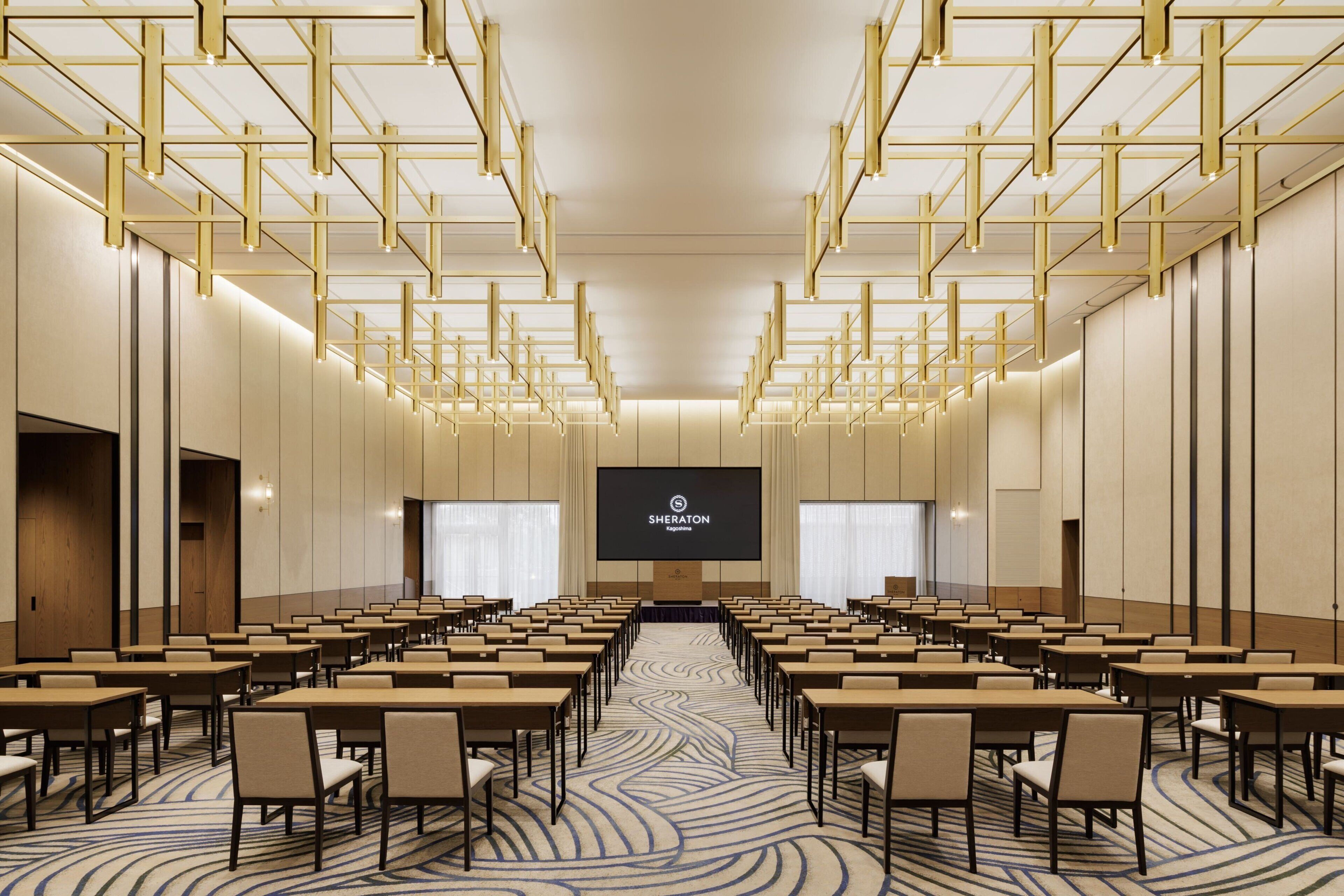 A conference room with rows of tables and chairs, a large screen at the front, and a patterned carpet.