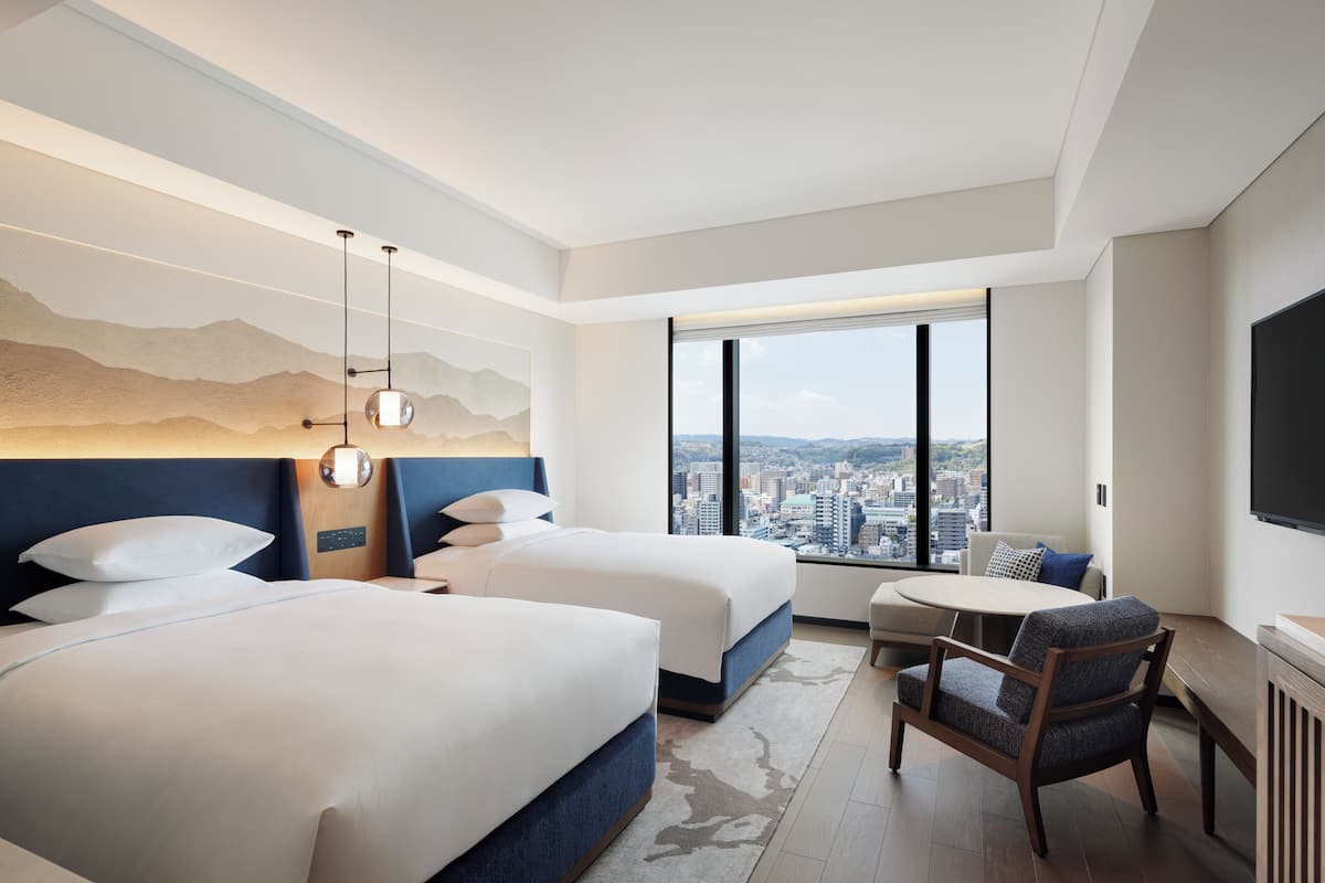 Ein Hotelzimmer mit zwei Betten, einer Sitzecke, einem großen Fenster mit Blick auf die Stadt und einem Flachbildfernseher.