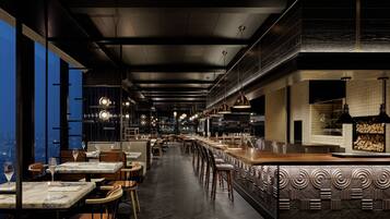 A modern restaurant with a long counter, high ceilings, and large windows overlooking a cityscape.