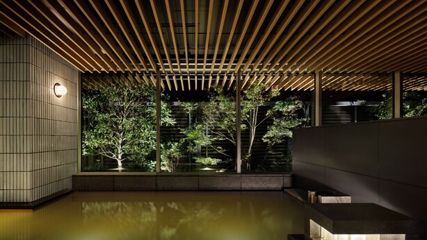 Un interior moderno con techo de madera, suelo reflectante y vista a un jardín con árboles y una fuente de agua.