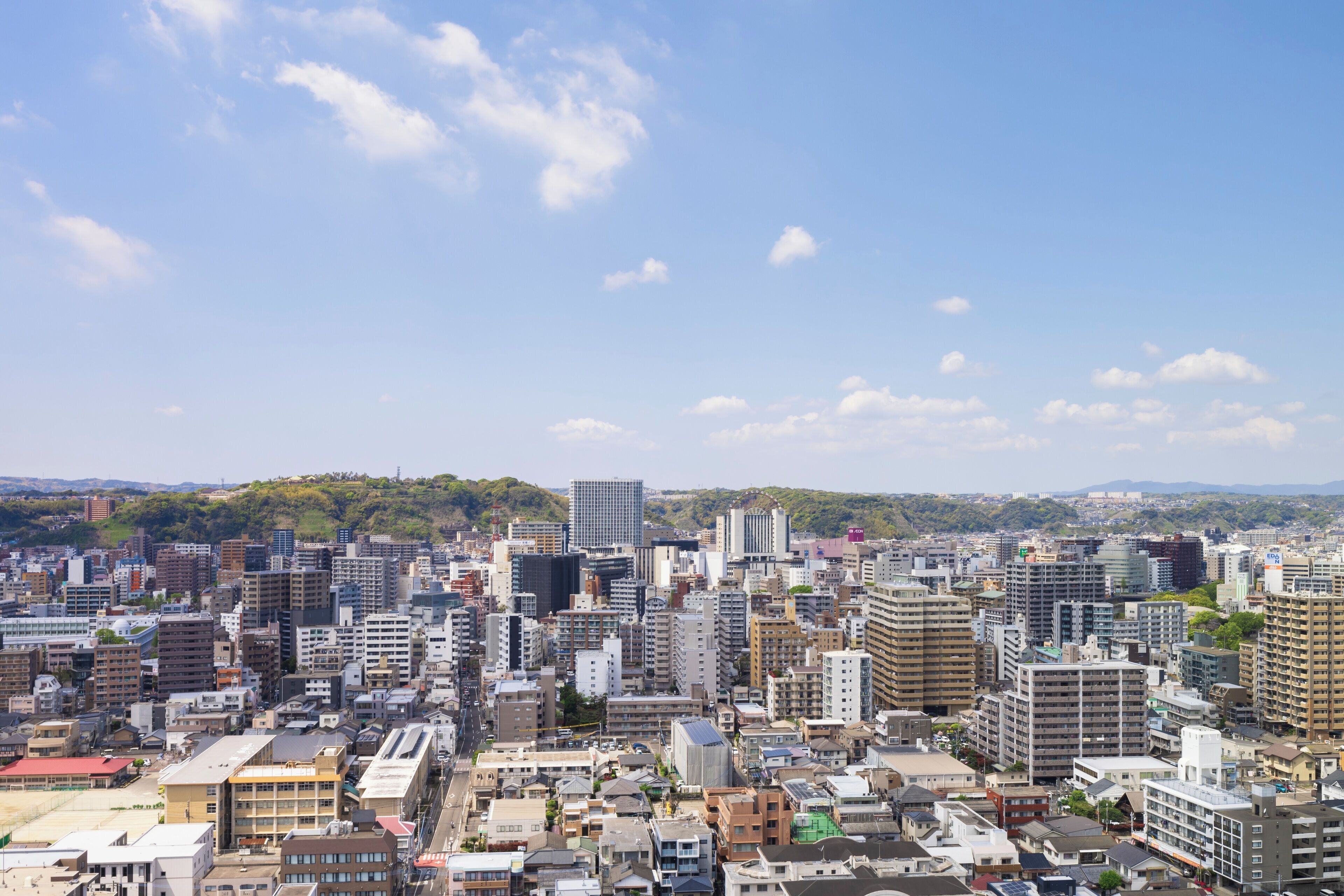 A cityscape with dense urban buildings, a clear sky, and distant hills.