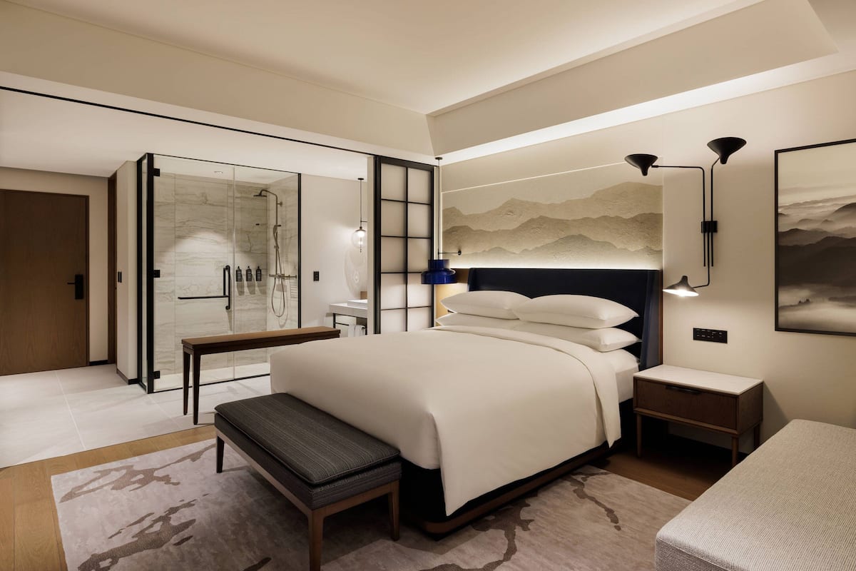 Ein modernes Hotelzimmer mit einem großen Bett, einer Bank, einem Nachttisch, einem Schreibtisch, einer Glasdusche und einer Wandleuchte.