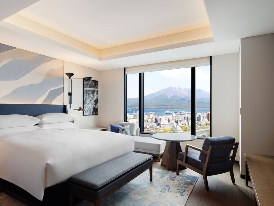 A hotel room with a large bed, a seating area, a small table, and a view of mountains and a city.