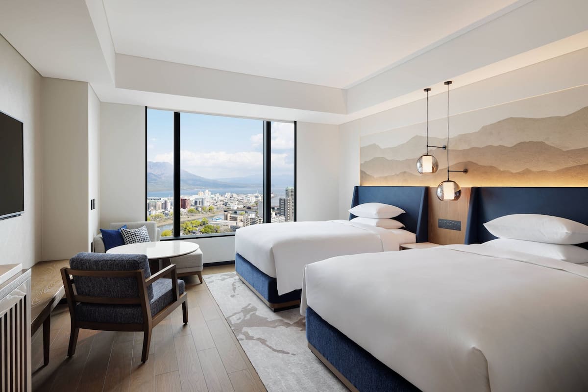 Ein Hotelzimmer mit zwei Betten, einem Sofa, einem Esstisch und einem großen Fenster mit Blick auf die Stadt.