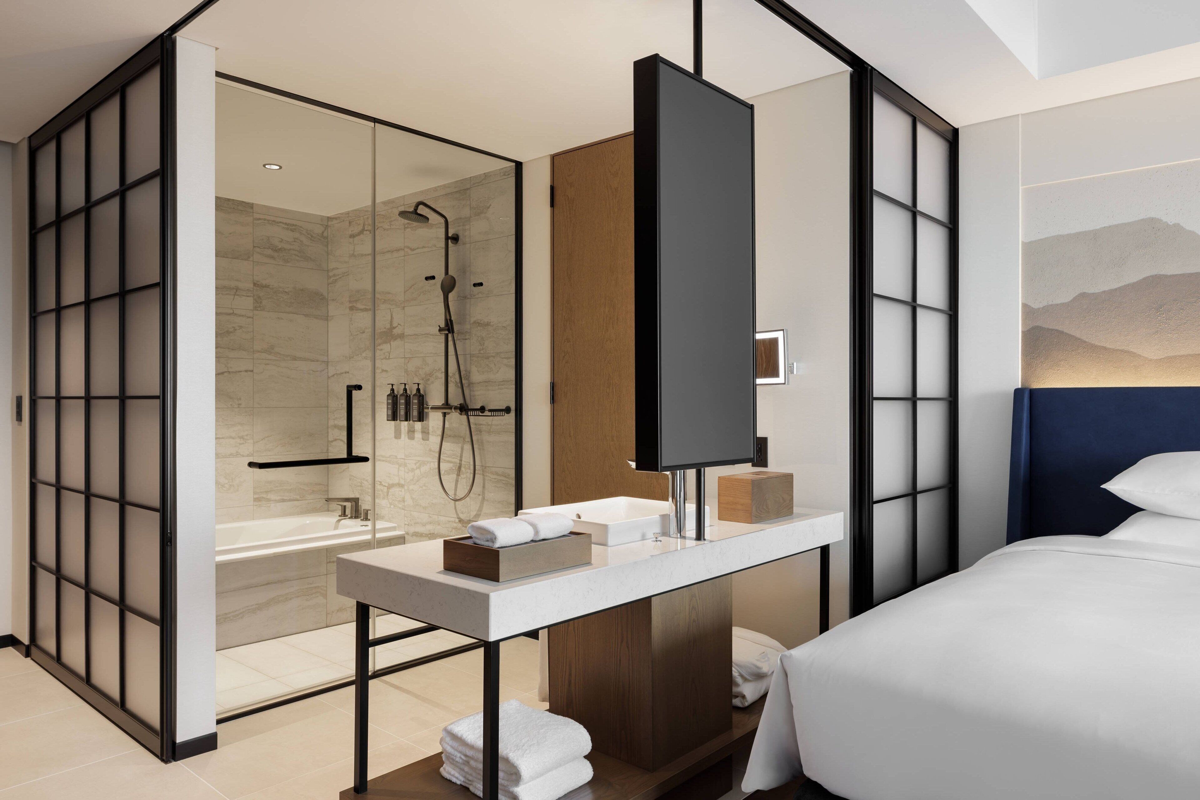 A modern hotel room with a glass-enclosed shower, a wooden vanity, a flat-screen TV, and a bed with white bedding.