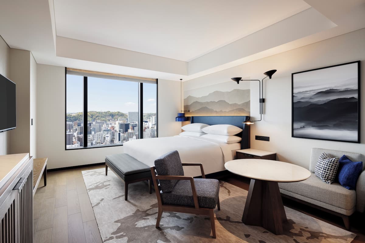 Ein modernes Hotelzimmer mit einem großen Bett, einem Schreibtisch, einem Sessel, einem runden Tisch und einer Couch. Ein großes Fenster bietet Blick auf die Stadt.