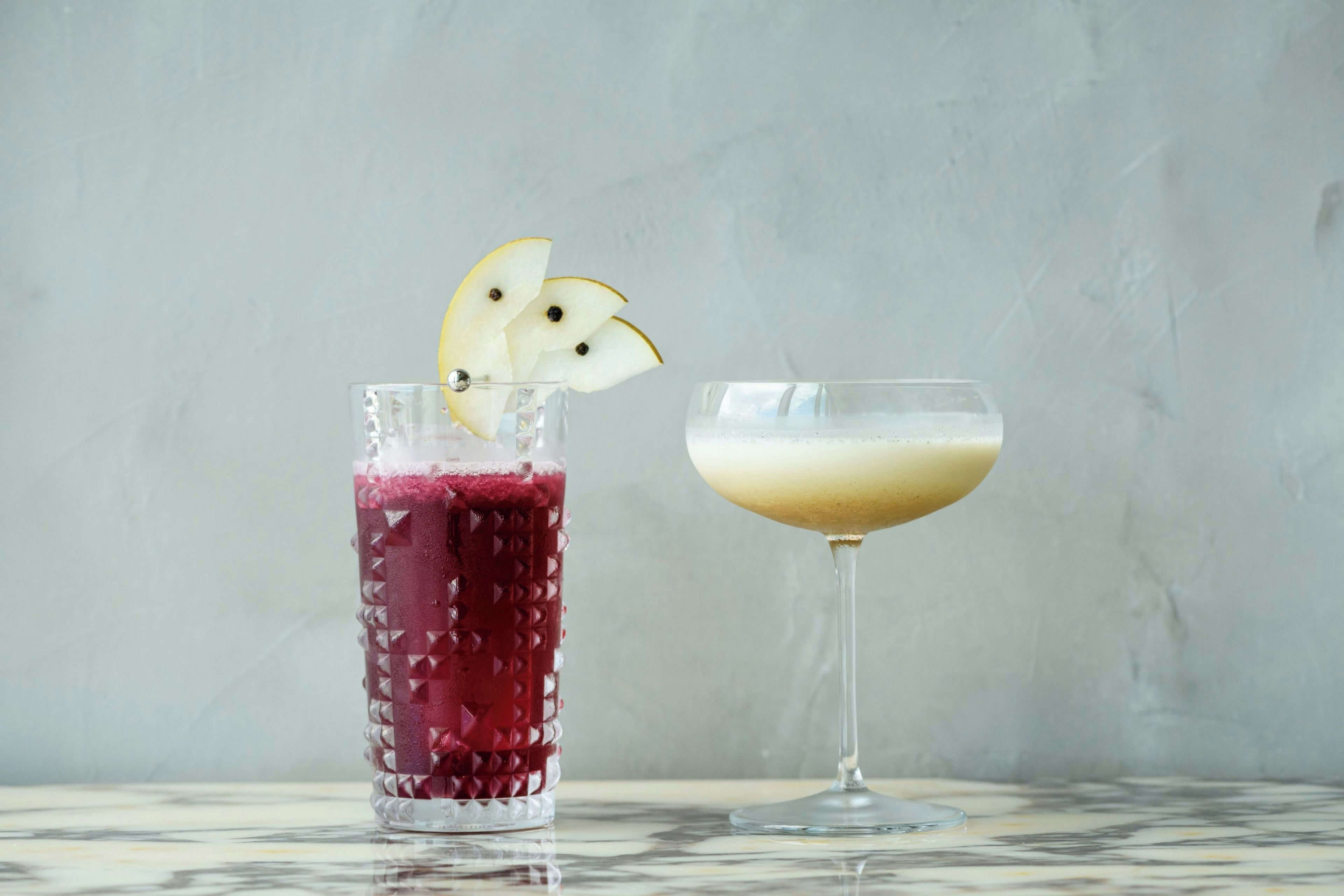 Dos vasos de bebidas, uno con un líquido rojo y otro con un líquido pálido, sobre una superficie de mármol.