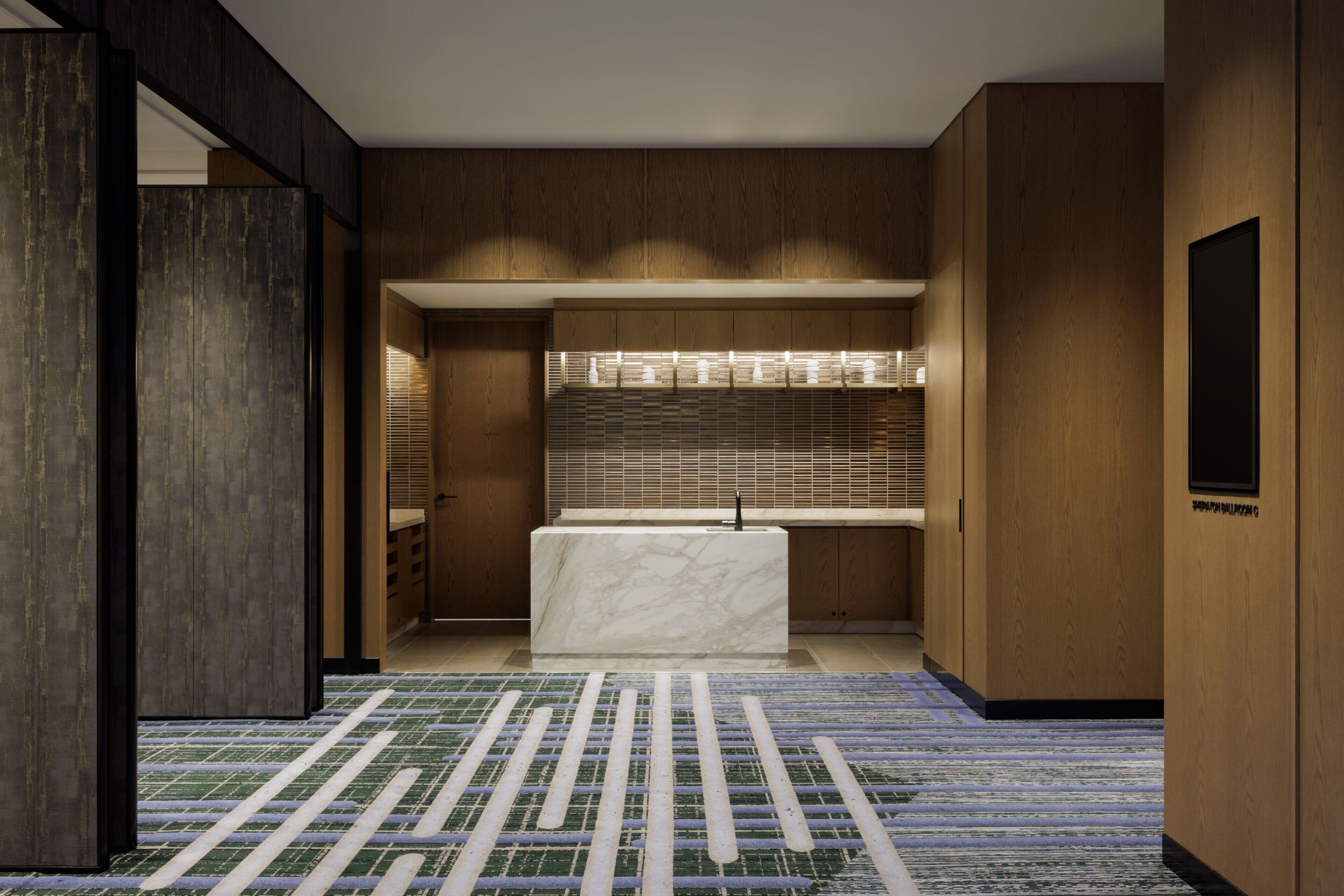 A modern hotel lobby with a marble reception desk, wooden paneling, and a patterned carpet.