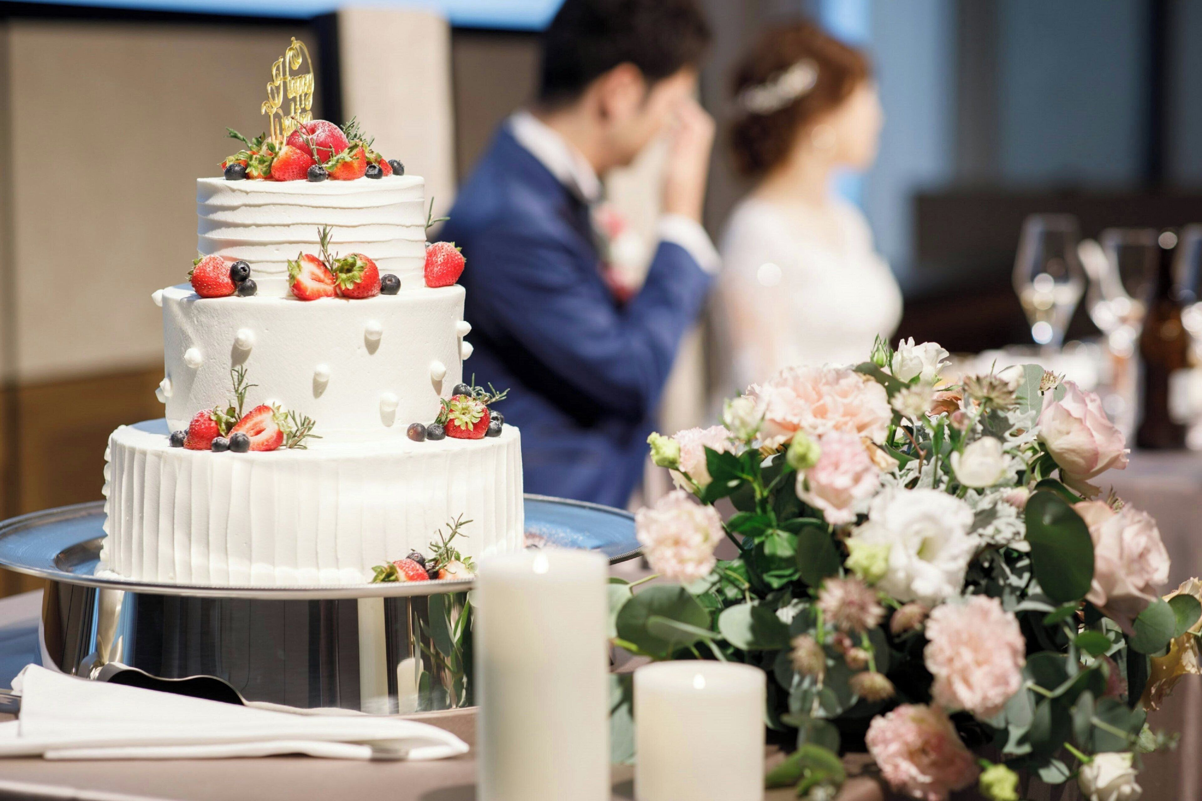 Un pastel de bodas con fresas y un adorno dorado, un arreglo floral y una vela sobre una mesa.