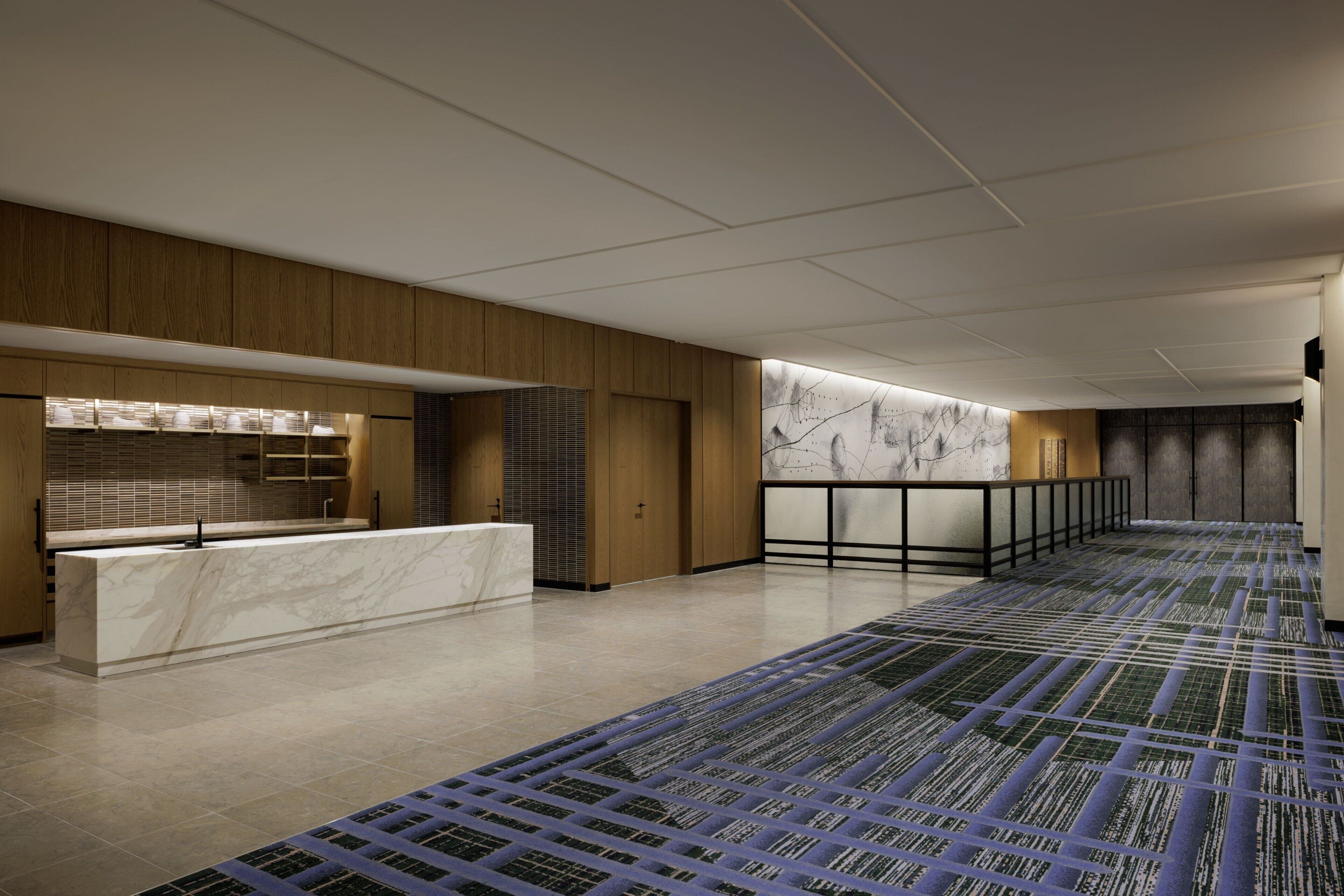 A modern hotel lobby with a marble reception desk, wooden paneling, a patterned carpet, and a large abstract wall mural.