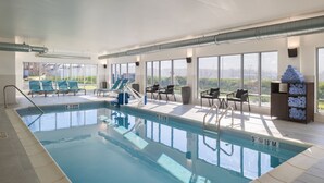 An indoor swimming pool with clear water, surrounded by sunlit seating areas and large windows offering views of the outdoors.