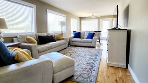 A spacious living room with a large sectional sofa, a smaller sofa, and a coffee table. There are two ceiling fans, a flat-screen TV, and a wooden floor.