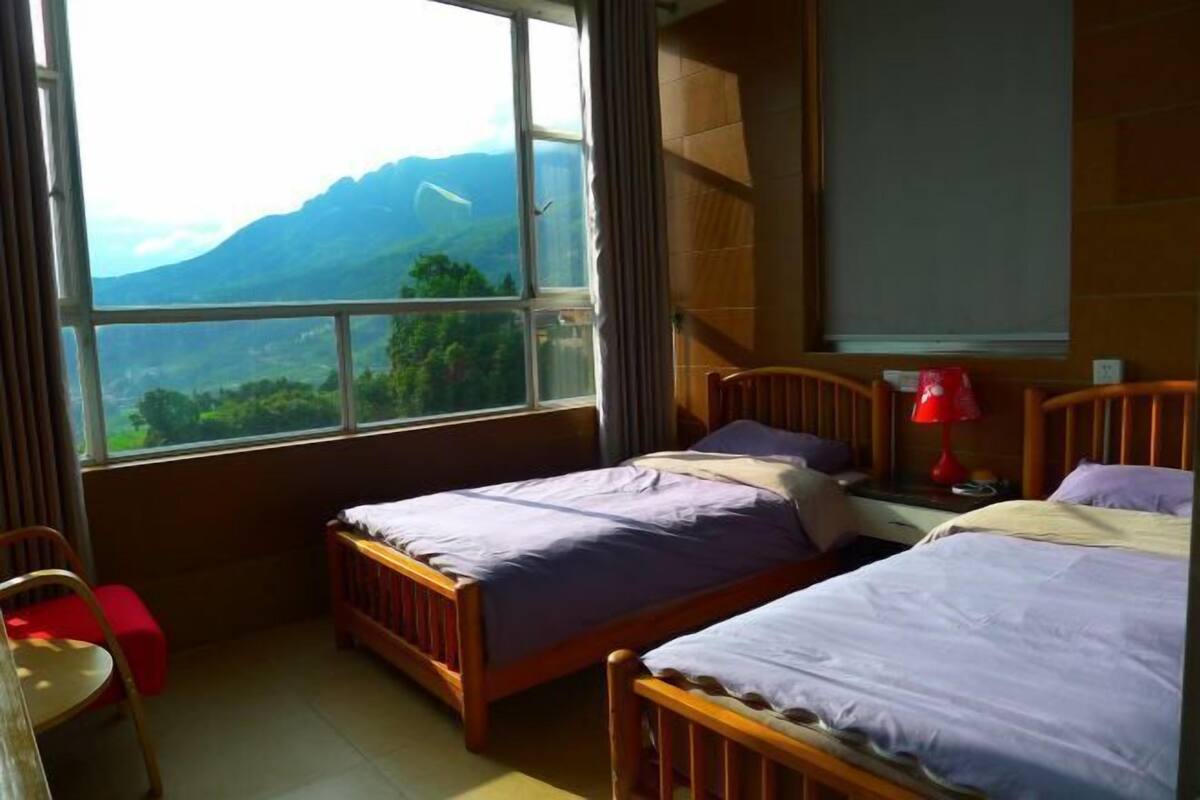 Ein Schlafzimmer mit zwei Betten, einem Fenster mit Bergblick, einem Sessel und einem Nachttisch mit Lampe.