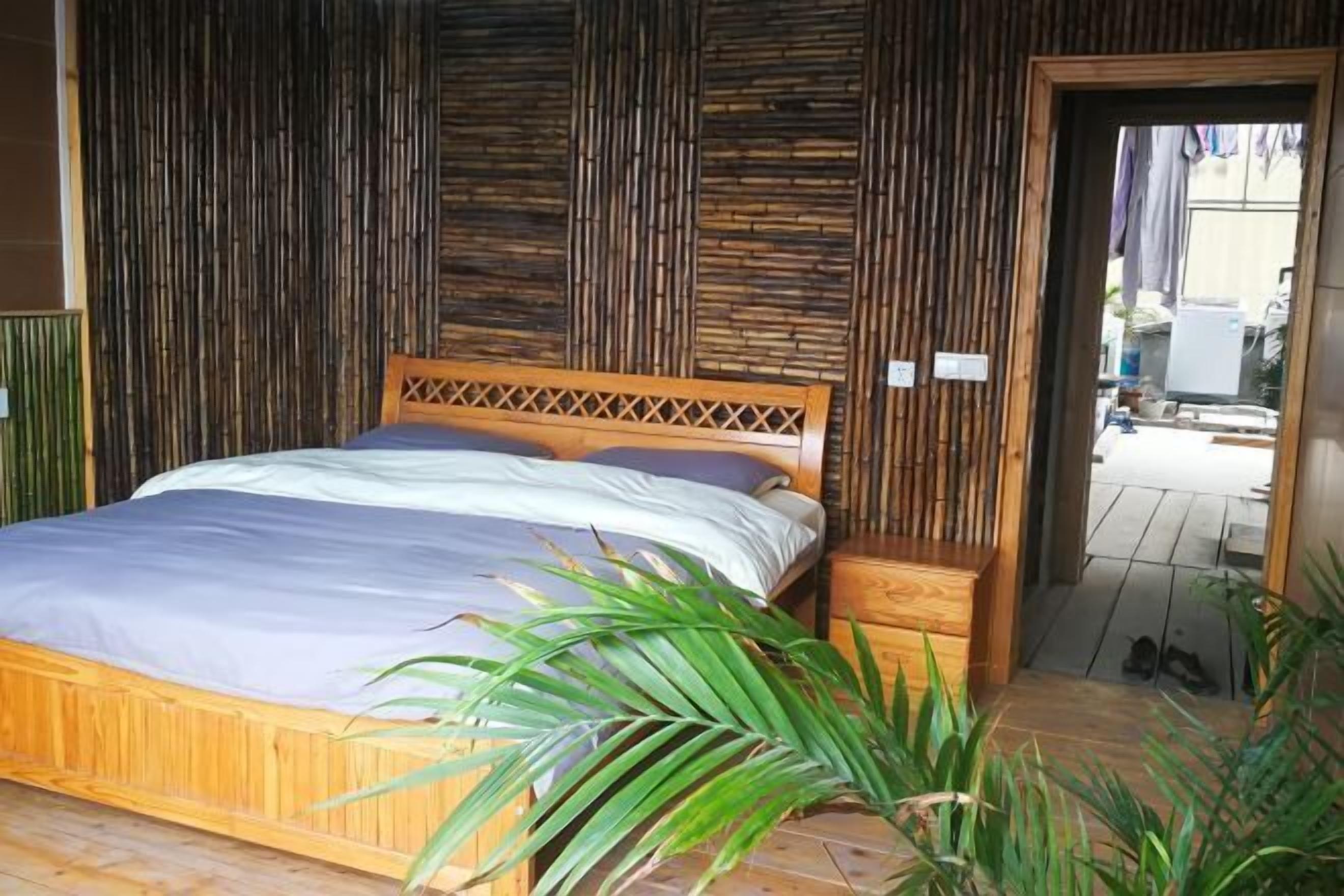 A wooden bed with a headboard, a bedside table, and a potted plant in a room with bamboo panel walls.