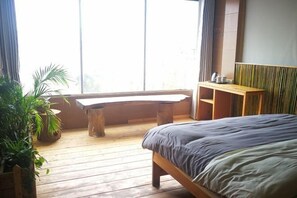 A bedroom with a wooden floor, a bed with a grey bedspread, a wooden bench, a small table with a cup and saucer, and a large window with curtains. - Yuanyang Green Hostel & Sunny Guesthouse (Yuanyang)