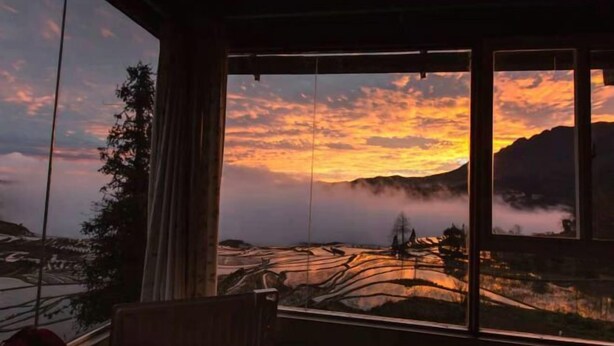 Een kamer met een groot raam met uitzicht op een landschap bij zonsondergang.