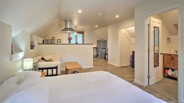 A spacious loft bedroom with a large bed, a small seating area, a kitchenette, and a view of the living area.