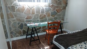 Une pièce avec un mur en pierre, une table en verre décorée d’autocollants, une chaise orange, un lit recouvert d’une couverture à motifs et un plancher en bois.