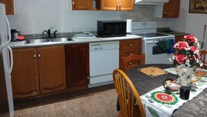Une cuisine avec des meubles de rangement en bois, un four à micro-ondes, une cuisinière, un lave-vaisselle et une table à manger recouverte d’une nappe.