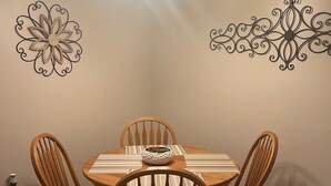Un comedor con una mesa redonda de madera, cuatro sillas de madera, un adorno en la pared y una araña.
