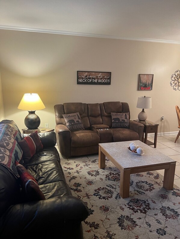 Ein Wohnzimmer mit einer braunen Ledersofa, einem Couchtisch, einem gemusterten Teppich und einem Schild an der Wand mit der Aufschrift "Welcome to our NECK OF THE WOODS".