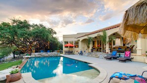 A swimming pool with lounge chairs and umbrellas, surrounded by a patio area with a thatched roof and outdoor seating. - Luxe Lake Travis Vacation Rental w/ Heated Pool (Lago Vista)