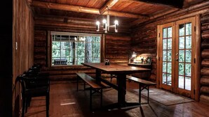 A rustic wooden cabin interior with a large wooden table and benches, a window with a view of greenery, and a door with glass panels.