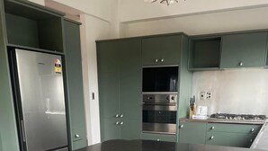 A modern kitchen with green cabinets, a stainless steel refrigerator, an oven, and a gas stove. There are two white stools in front of the counter. - Stylish CBD apartment with stunning river views (Hamilton)