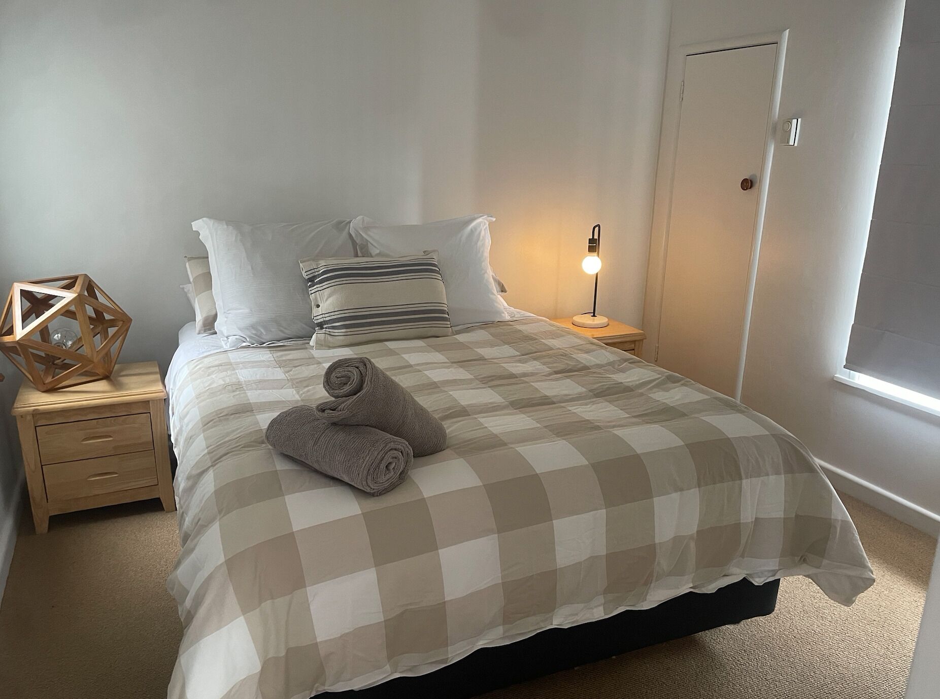 Un lit bien fait, avec une couvre-lit à carreaux, deux oreillers, une lampe de chevet et un objet géométrique en bois posé sur la table de chevet.