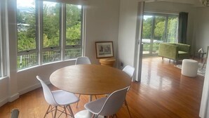 A spacious room with a wooden dining table and white chairs, large windows with a view of greenery, and a green sofa in the background. - Stylish CBD apartment with stunning river views (Hamilton)