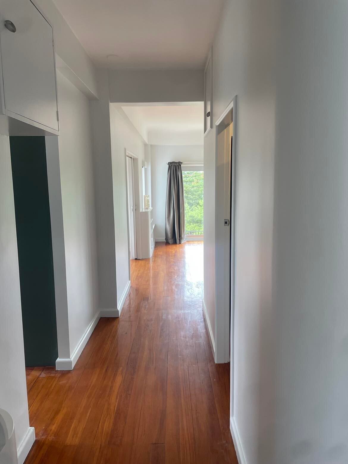 A hallway with wooden flooring, white walls, and a view of a garden through a window with a grey curtain.
