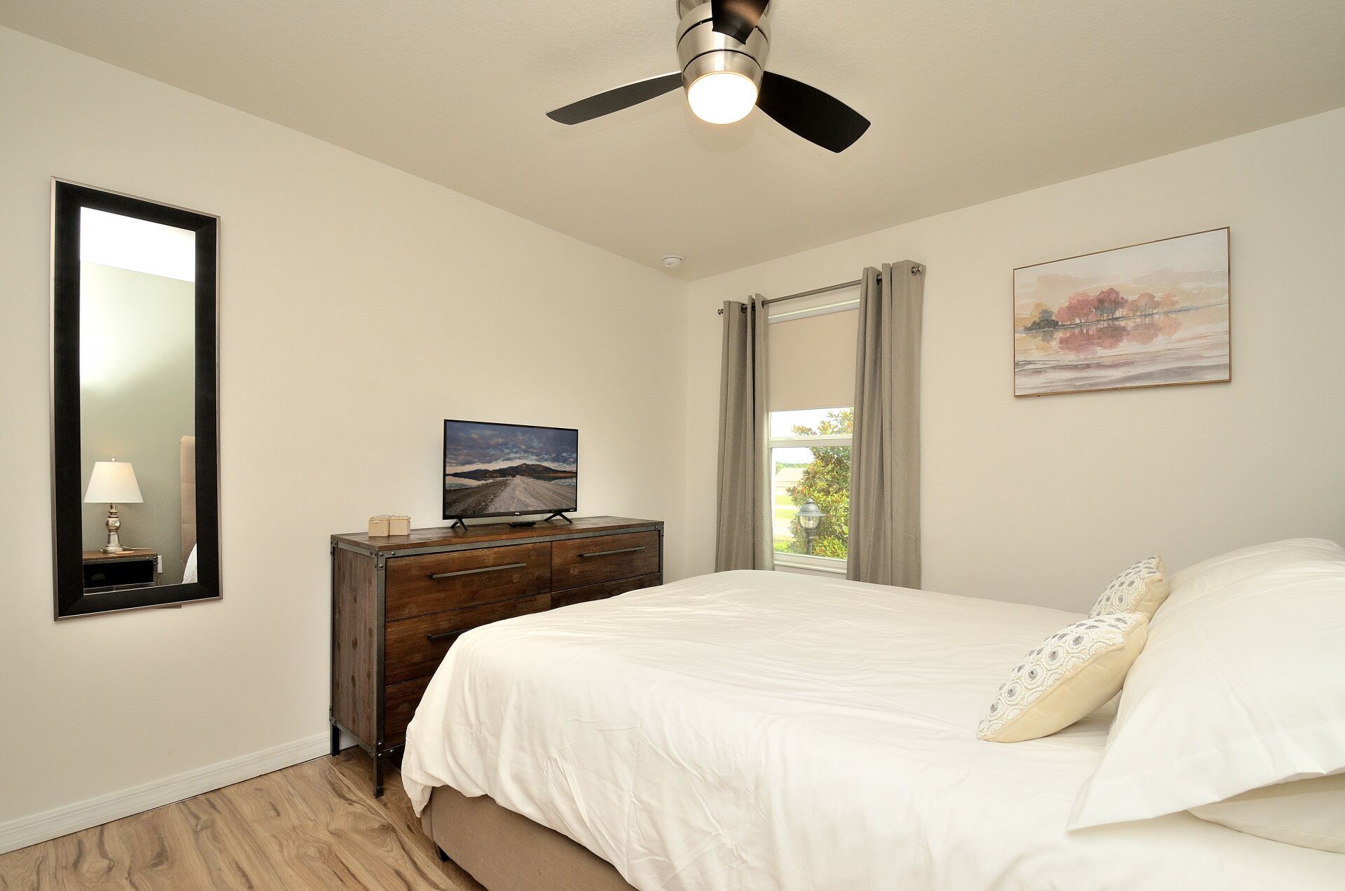 Een slaapkamer met een bed, een dressoir, een televisie, een plafondventilator, een raam met gordijnen en een schilderij aan de muur.
