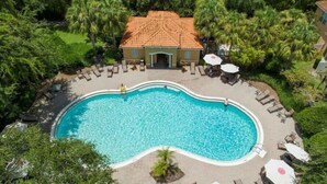 Eine Vogelperspektive zeigt einen Swimmingpool, umgeben von Liegestühlen und Sonnenschirmen, mit einem Gebäude im Hintergrund.