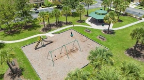 An aerial view of a park with a playground, including swings and a slide, surrounded by palm trees and a gazebo.
