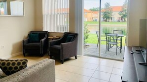 Un vaste espace de vie comprenant un canapé, deux fauteuils, une table basse et un téléviseur. Une porte-fenêtre coulissante donne accès à un patio extérieur équipé d’une table et de chaises.